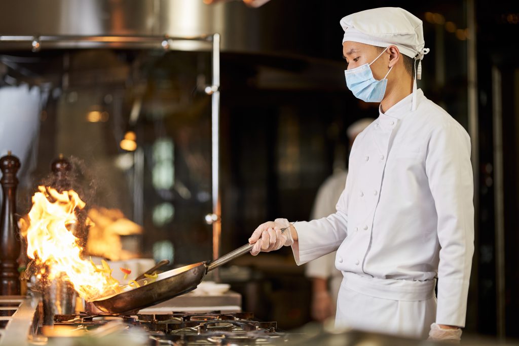 Concentrated chef in face mask holding a flaming pan over a stove while preparing a meal