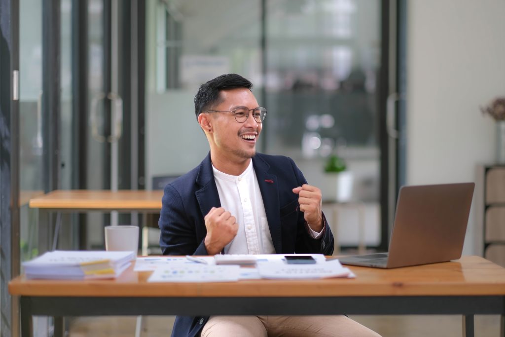 Portrait of an Asian male business owner standing with a computer showing happiness after a successful investment.